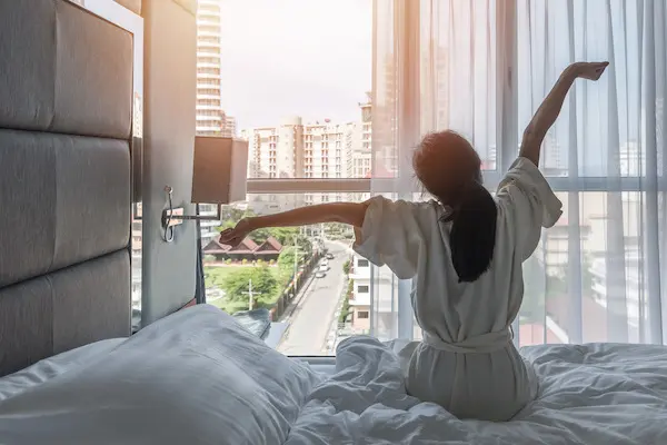 Woman in a white robe sitting on a hotel bed looking out of the hotel window and stretching as if she just woke up